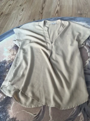 Beige blus från Bubbleroom - Stilren beige blus från Bubbleroom med korta ärmar och v-ringning. Blusen har en avslappnad passform och är tillverkad i ett mjukt, lätt material som ger en skön känsla. Perfekt att matcha med jeans eller kjol för en trendig look.