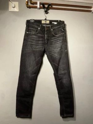 Svarta slim fit jeans från Replay - Säljer ett par svarta slim fit jeans från Replay med snyggt slitna detaljer och klassisk femficksmodell. Jeansen har en något tvättad look och raka ben. Perfekta för dig som gillar en modern och avslappnad stil.