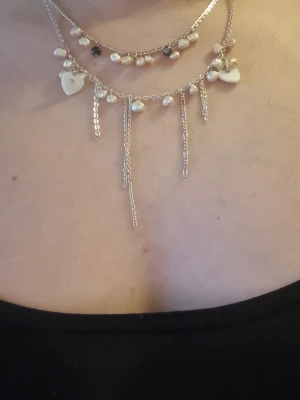 Silverfärgat halsband med pärlor och hjärtan - Snyggt lager-på-lager halsband i silverfärg med små vita pärlor, svarta detaljer och flera hängande kedjor. Halsbandet har även söta hjärtformade berlocker som ger en romantisk touch. Perfekt accessoar för att lyfta din outfit.