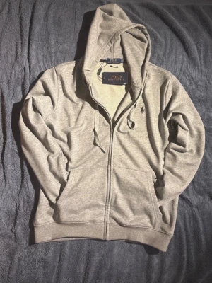 Grå hoodie från Polo Ralph Lauren - Snygg grå hoodie från Polo Ralph Lauren med dragkedja, huva med dragsko och klassisk broderad logga på bröstet. Tillverkad i mjukt bomullsmaterial och har två fickor framtill. Perfekt för en chill och stilren look.