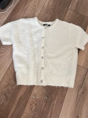 Vit fluffig kofta från Gina Tricot - Kort, vit kofta från Gina Tricot i fluffigt material. Koftan har rund halsringning, korta ärmar och knäpps med silvriga knappar framtill. Perfekt för att addera en mysig touch till din outfit.