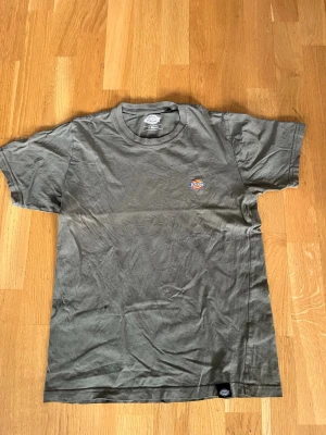 Grå Dickies t-shirt med liten logga - Säljer en grå t-shirt från Dickies med liten broderad logga på bröstet. Klassisk rund hals och korta ärmar. Perfekt basic-plagg för en avslappnad stil. Tillverkad i mjuk bomull som känns skön mot huden.