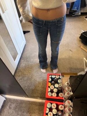 Blå bootcut jeans med hål - Säljer ett par blå bootcut jeans med hål på låret och knäna och snygga detaljerade fickor fram. Jeansen har låg midja och klassisk femficksdesign. Perfekta för dig som gillar en avslappnad men trendig look. S barn.