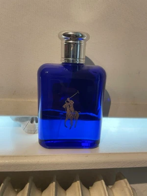 Ralph Lauren Polo Blue parfym - En snygg parfymflaska från Ralph Lauren i djupblått glas med silverfärgat lock. Flaskan har den klassiska Polo-loggan med en ryttare i silver på framsidan och RL ingraverat på locket. Perfekt för dig som gillar stilrena och exklusiva dofter. 40 ml kvar