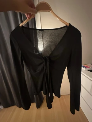Svart långärmad topp med knyt - Trendig svart långärmad topp med djup v-ringning och snygg knytning framtill. Perfekt att styla med både jeans och kjol för en cool look. Tunn och mjuk i materialet, vilket gör den skön att bära hela dagen.