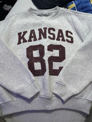 Grå sweatshirt med Kansas-tryck - Säljer en grå sweatshirt  med stort vinrött tryck 'KANSAS 82' på bröstet. Tröjan har rund halsringning, ribbade muddar vid ärmslut och nederkant. Perfekt för dig som gillar college-stil och vill ha en skön och avslappnad look.