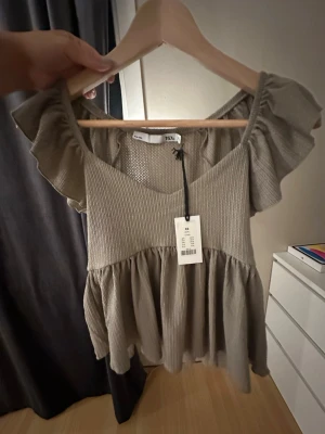 Beige volangblus från 157 - Supersöt beige topp från 157 med volangärmar och v-ringning. Toppen har en luftig, flowy passform och ribbad struktur upptill. Perfekt för dig som gillar en avslappnad men ändå trendig stil. Lappen sitter kvar.