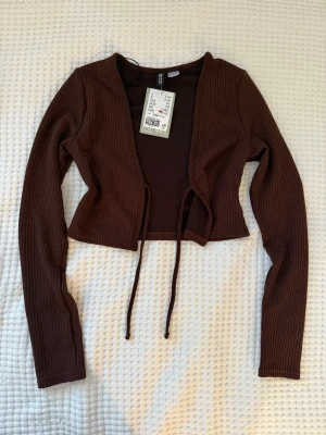 Brun ribbad croppad bolerotopp Gina Tricot - Snygg brun croppad bolerotopp från Gina Tricot med långa ärmar och knyt framtill. Toppen är ribbad och har en öppen design som ger en trendig look. Perfekt att styla över ett linne eller topp för en cool layering-effekt.