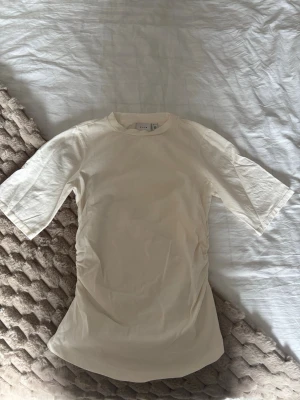 Offwhite t-shirt med rynkade sidor - Säljer en offwhite t-shirt från Vila med rynkade detaljer på sidorna. T-shirten har rund halsringning och lite längre ärmar än vanligt. Tillverkad i mjuk bomull som känns skön mot huden. Perfekt för en clean och stilren look.