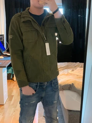 Olivgrön vindjacka med dragkedja - Snygg olivgrön vindjacka från GUILD WARS med dragkedja framtill och flera praktiska fickor. Jackan har klassisk krage och lång ärm, perfekt för dig som vill ha en stilren och funktionell look. Passar till jeans och t-shirt för en avslappnad vibe.