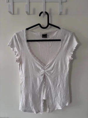 Vit v-ringad topp från Gina  - Söt vit topp från Gina Tricot med djup v-ringning och ”skärning” framtill. Kortärmad modell med vågiga kanter och lätt croppad passform. Tillverkad i mjuk bomull, perfekt för en avslappnad och trendig look.