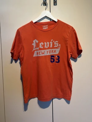 Orange Levi's t-shirt med tryck - Säljer en orange t-shirt från Levi's med coolt New York-tryck och siffran 53 framtill. T-shirten är i 100% bomull och har korta ärmar. Perfekt för dig som gillar streetwear och vill ha något bekvämt och stilrent.