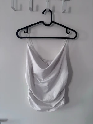 Vit draperad linnetopp med tunna band - Säljer en stilren vit linnetopp med tunna axelband och snygg drapering framtill. Toppen har en croppad passform och är perfekt för att skapa en trendig look. Tillverkad i mjukt material som känns skönt mot huden.