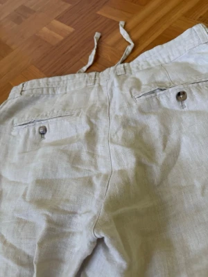 Beiga linnebyxor med dragsko - Snygga och luftiga linnebyxor i beige med dragsko i midjan och knäppning fram. Byxorna har två sidofickor och två bakfickor med knapp. Perfekta för varma dagar och en avslappnad stil.