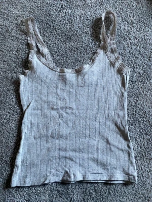 Ljusgrå ribbad linnetopp med spets - Ljusgrå linnetopp med ribbad struktur och fina spetsdetaljer längs axelbanden och halsringningen. Toppen är ärmlös och har en slim passform som framhäver siluetten. Perfekt för att skapa en avslappnad och trendig look.