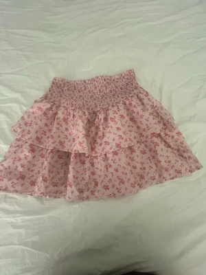 Rosa blommig volangkjol - Supersöt kort kjol i ljusrosa med små blommor och bred smock i midjan. Kjolen har dubbla volanger som ger en fluffig och lekfull look. Perfekt för dig som gillar romantisk och feminin stil.