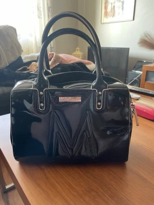 Svart lackad handväska från Mariamare - Snygg svart handväska i glansigt lackmaterial från Mariamare. Väskan har två rejäla handtag, silverfärgade metalldetaljer och ett stort M framtill. Praktisk dragkedja på baksidan och rymlig insida. Perfekt för dig som gillar statement-accessoarer. Bild visar även renlighet inne i väskan där den tydligt är näst intill oanvänd.