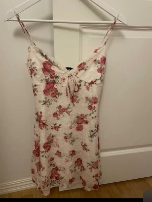 Blommig spetsklänning från Bershka - Söt kort klänning från Bershka i ljus spets med blommigt mönster i rosa och grönt. Klänningen har smala axelband, liten rosett framtill och figurnära passform. Perfekt för sommarens alla tillfällen.