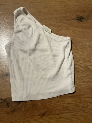 Vit oneshoulder topp från Even&Odd - Säljer en ribbad vit oneshoulder topp från Even&Odd. Toppen är croppad och har en snygg asymmetrisk axelbandslös design som ger en trendig look. Perfekt att matcha med höga jeans eller kjol för en cool outfit.