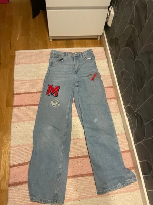 Blåa jeans - Ett par blåa jeans med ett rött M på benet. Den har ett litet hål på knät och trycket är väldigt urtvättat. 