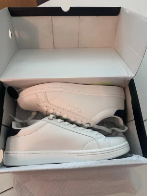 Vita sneakers från Hush Puppies - Stilrena vita sneakers från Hush Puppies med lågt skaft och klassisk design. Skorna har vita snören, diskret logga på sidan och svart detalj vid hälen. Tillverkade i slätt syntetmaterial som ger en clean och fräsch look.