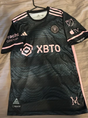 Inter Miami Messi matchtröja svart/rosa - Snygg svart Inter Miami CF matchtröja från Adidas med rosa detaljer och Messi 10-tryck på ryggen. Tröjan har klubbmärke, sponsorloggor och MLS-märke på ärmen. Tillverkad i lätt funktionsmaterial med Aeroready-teknologi för bra ventilation.