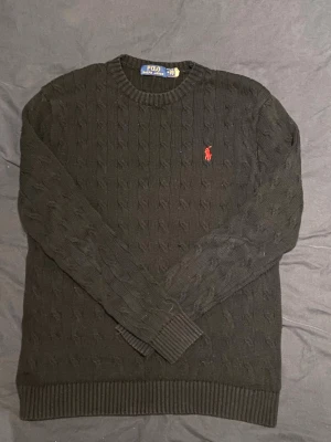 Mörkgrön kabelstickad tröja Polo Ralph Lauren - Kabelstickad svart tröja från Polo Ralph Lauren med rund halsringning och klassisk röd logga broderad på bröstet. Tröjan har ribbade muddar vid ärmslut och nederkant. Perfekt för lager på lager och en stilren look. Väldigt skön och lätt att styla till,kom med frågor och funderingar, pris kan dessutom diskuteras 