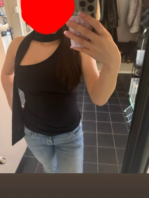 Svart oneshoulder topp med scarfdetalj - Cool svart oneshoulder topp med scarfdetalj runt halsen och öppen rygg. Toppens asymmetriska design ger en edgy look och den är tight i passformen. Perfekt att styla med jeans eller kjol för en trendig outfit.