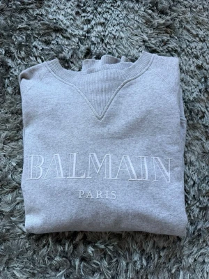 Grå sweatshirt från Balmain Paris - Säljer en stilren grå sweatshirt från Balmain Paris med broderad logga framtill. Tröjan har rund halsringning, ribbade muddar vid ärmslut och nederkant samt en klassisk relaxed fit. Perfekt för dig som gillar exklusiva streetwear-vibbar.