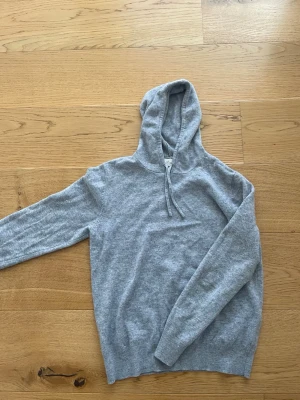 Grå cashmere hoodie från Sayless - Mjuk och stilren hoodie i ljusgrått från Sayless, tillverkad i lyxig cashmere. Klassisk modell med huva och dragsko, perfekt för en chill och avslappnad look. Långa ärmar och ribbade muddar ger en skön passform.