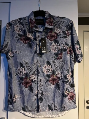 Blommig skjorta från Replay - Snygg kortärmad skjorta från Replay med blommigt mönster i vinrött, vitt och grönt på en blå bas. Skjortan har klassisk krage och knappar framtill, perfekt för en avslappnad och trendig look.