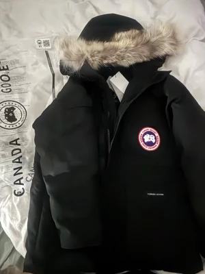 Svart dunjacka från Canada Goose - Säljer en svart dunjacka från Canada Goose med Fusion Fit. Jackan har en stor huva med äkta päls, klassisk logga på bröstet och dragkedja framtill. Perfekt för kalla vinterdagar och riktigt varm. Snygg och tidlös design. Jag säljer den för att den va för stor, XS är den men passar nog mer en S-M. 