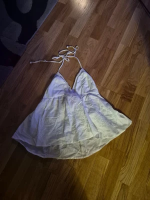 Vit broderad linnetopp med knyt - Söt vit linnetopp med broderade detaljer och tunna axelband som knyts i nacken. Toppen har v-ringning och är tillverkad i lätt bomull, perfekt för varma dagar. Fladdrig passform och romantisk känsla.