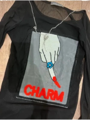 Svart mesh topp med CHARM-tryck - Cool svart mesh topp från Vince med lång ärm och ett stort tryck framtill av en hand med blå ring och röd texten 'CHARM'. Toppen är transparent och har en edgy vibe, perfekt för att sticka ut.