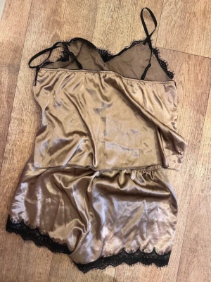 Champagnefärgat satinset med spets - Snyggt tvådelat set i champagnefärgad satin med svarta spetsdetaljer. Setet består av ett linne med tunna axelband och V-ringning samt matchande shorts med elastisk midja. Båda delarna har dekorativ svart spets längs kanterna för en extra lyxig känsla.