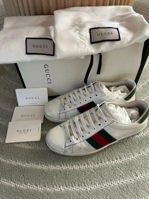 Gucci vita sneakers med ränder - Säljer ett par klassiska vita sneakers från Gucci i läder med de ikoniska gröna och röda sidoremsorna. Skorna har lågt skaft, vita snören och diskret logga på hälen. Perfekta för dig som vill ha en stilren och lyxig look.