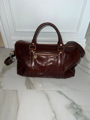 Mörkbrun weekendbag i läder *Aldrig använd*  - Snygg mörkbrun weekendbag i äkta läder med guldfärgade metalldetaljer. Väskan har både handtag och justerbar axelrem samt en praktisk ytterficka med dragkedja. Perfekt för resor eller övernattningar när du vill ha stil och funktion.