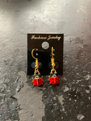 Guldörhängen med nyckelpigor - Söta örhängen i guldton med hängande röda nyckelpigor dekorerade med svarta prickar. Krokarna är guldfärgade och nyckelpigorna är detaljerade i rött och svart. Perfekt för dig som vill ha något lekfullt och unikt i din stil.