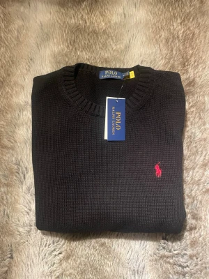 Svart stickad tröja Polo Ralph Lauren - Svart stickad tröja från Polo Ralph Lauren med rund halsringning och klassisk röd broderad logga på bröstet. Tröjan har ribbstickade muddar och är tillverkad i ett mjukt material som passar perfekt till säsongens outfits.