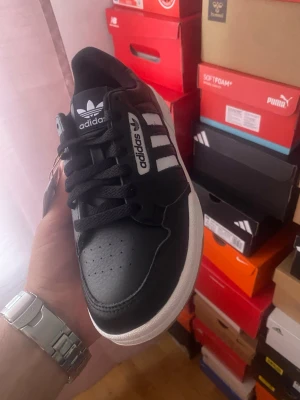 Svarta Adidas sneakers med vita ränder - Klassiska svarta Adidas sneakers med vita ränder och vit sula. Skon har lågt skaft, svart ovandel i läder och tydliga Adidas-loggor på plös och sida. Snygg retrostil som passar till många outfits.
