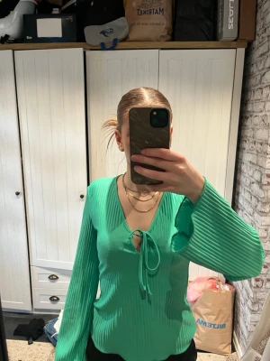Grön ribbad blus med knyt - Trendig grön blus med ribbad struktur och långa ärmar. Djup v-ringning med snygg knytning framtill. Perfekt för dig som vill ha en färgklick i garderoben och gillar en relaxed men ändå chic stil.