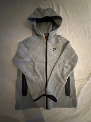 Grå zip hoodie från Nike - Snygg grå hoodie från Nike med dragkedja framtill och svart Nike-logga på bröstet. Hoodien har två svarta dragkedjeförsedda fickor och en stor huva. Har knappt använt den då den är i väldigt bra skick. Hör av dig vit mer frågor och information. 