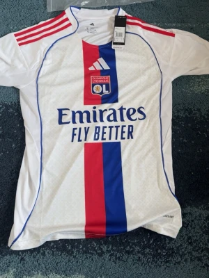 Olympique Lyonnais matchtröja Adidas - Säljer en vit Olympique Lyonnais matchtröja från Adidas med röda och blå detaljer, klubbmärke på bröstet och Emirates Fly Better-tryck. Tröjan har röda ränder på axlarna och är tillverkad i ett lätt, ventilerande material.