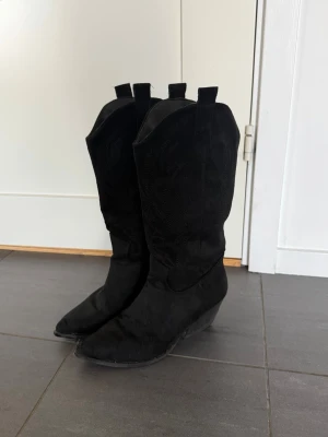 Svarta cowboy boots med kilklack - Snygga svarta cowboy boots med spetsig tå och kilklack. Boots i mockaimitation med klassiska western-detaljer och dragflikar upptill. Perfekta för dig som vill ha en cool och tidlös look.