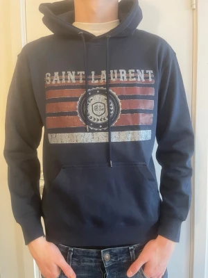 Mörkblå Saint Laurent hoodie - Snygg mörkblå hoodie från Saint Laurent med tryck i vitt och rött framtill. Klassisk känguruficka och justerbar huva med snören. Perfekt för dig som vill ha en streetwear-look med exklusiv känsla. Tillverkad i mjuk bomullsblandning.        Bröstomfång: 98 cm Axelbredd: 44 cm Plagglängd: 63 cm Ärmlängd: 56 cm