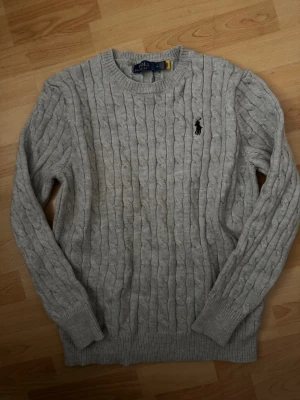 Grå kabelstickad tröja Polo Ralph Lauren - Säljer en grå kabelstickad tröja från Polo Ralph Lauren med klassisk rund halsringning och svart broderad logga på bröstet. Den har ett hål i en armhålan som man kan se i sista bilden men annars i väldigt bra skick