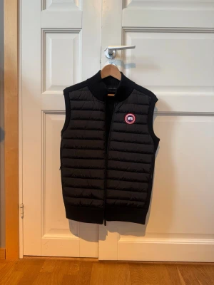 Svart dunväst från Canada Goose - Snygg svart dunväst från Canada Goose med ribbad krage och dragkedja framtill. Västen har det klassiska runda Canada Goose-märket på bröstet och quiltad framsida. Perfekt för lager-på-lager och håller dig varm under kyliga dagar. Andvänd väldigt fåtal gånger och är i nyskick. Köpt på NK i Göteborg, nypris: 6149kr, kvitto följer med. Skriv vid minsta fundering! Pris kan diskuteras 