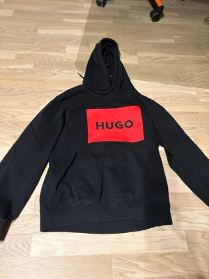 Svart hoodie från HUGO med röd logga - Snygg svart hoodie från HUGO med stor röd logga på bröstet och svart text. Klassisk huva med dragsko och känguruficka framtill. Tillverkad i mjukt material som känns skönt mot huden. Perfekt för dig som gillar streetwear och vill sticka ut.