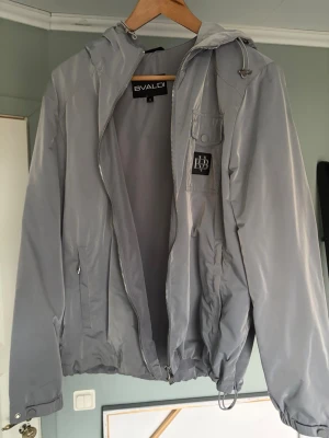 Grå vindjacka från Bvaldi - Snygg grå vindjacka från Bvaldi med huva och dragkedja framtill. Jackan har en ficka med lock och tryckknapp på bröstet samt en svart patch med Bvaldi-logga. Resår i midja och ärmslut. Använt den 1 gång och nu har den blivit för liten! Påse tillkommer. Pris är inte hugget i sten!