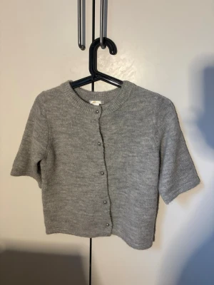Grå kortärmad kofta med knappar - Mysig grå kofta med korta ärmar och rund halsringning. Koftan har en rak passform och stängs med små knappar framtill. Perfekt att slänga över en topp för en enkel och stilren look.